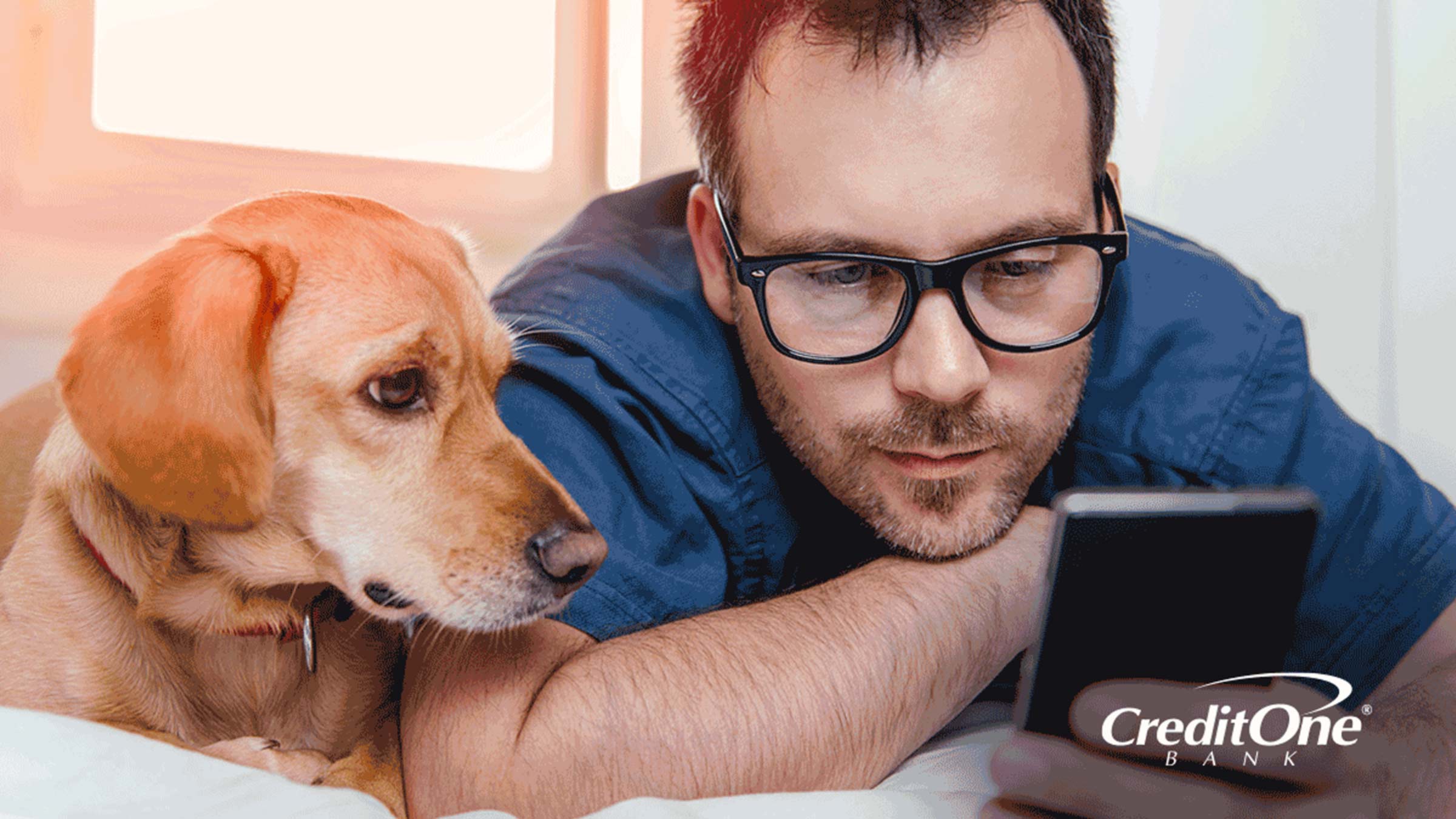 A man and his dog lie flat on his bed, both glancing at his smartphone. He looks relaxed, possibly enjoying the benefits of organizing his life after learning how to automate his finances.