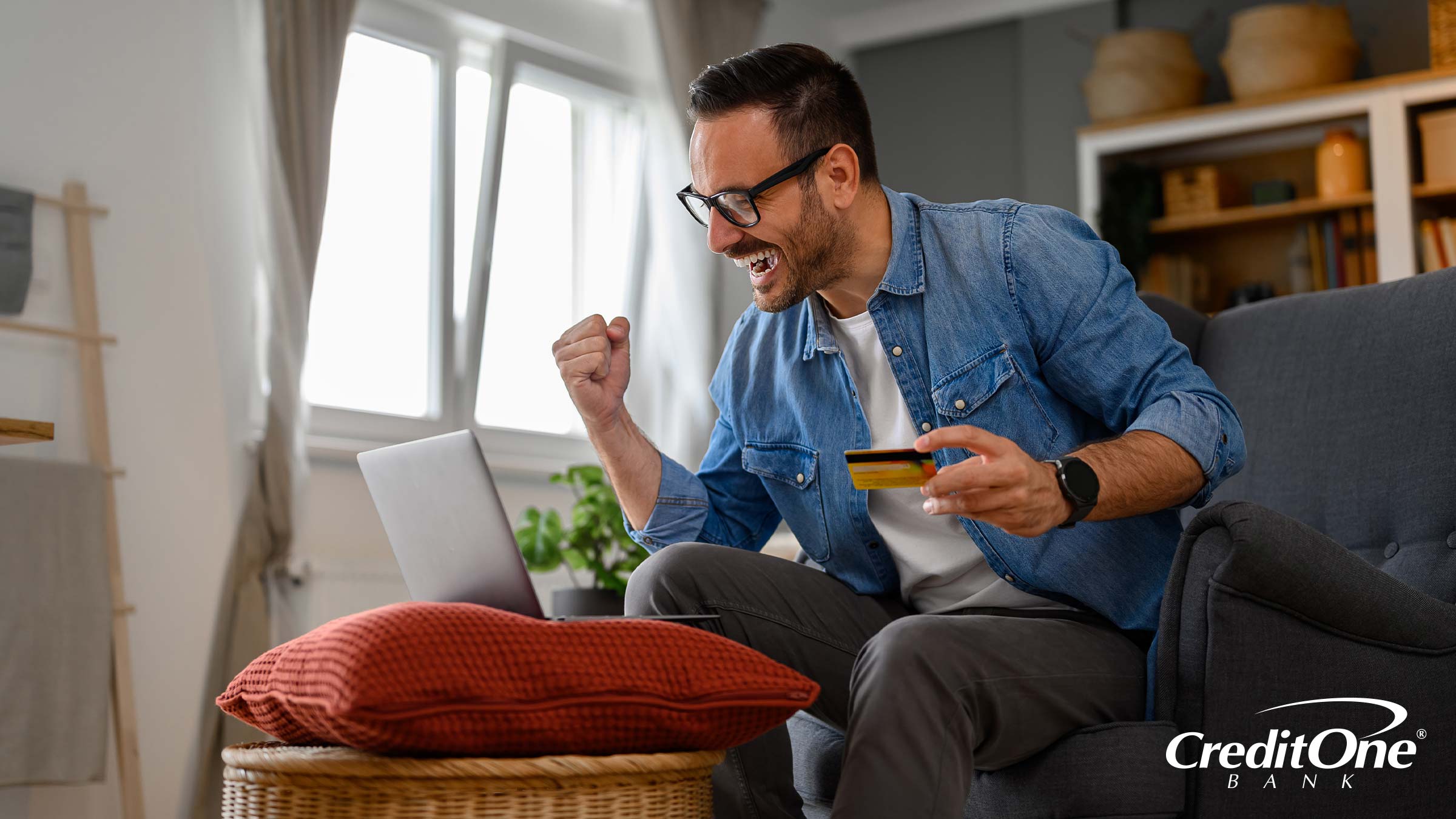 A man celebrates in front of his laptop with a credit card in hand. Perhaps he made a purchase and is experiencing firsthand how credit card cash back works.