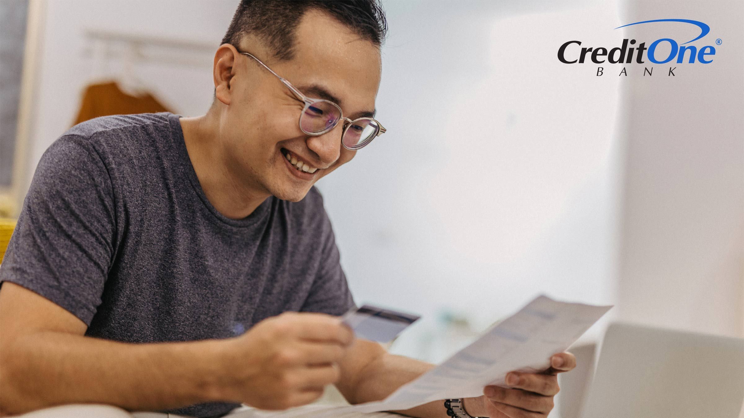A man holds a credit card in one hand as he reviews a document he’s holding in the other. He is smiling, perhaps because this is the first step he’s taking toward rebuilding his credit with credit cards.