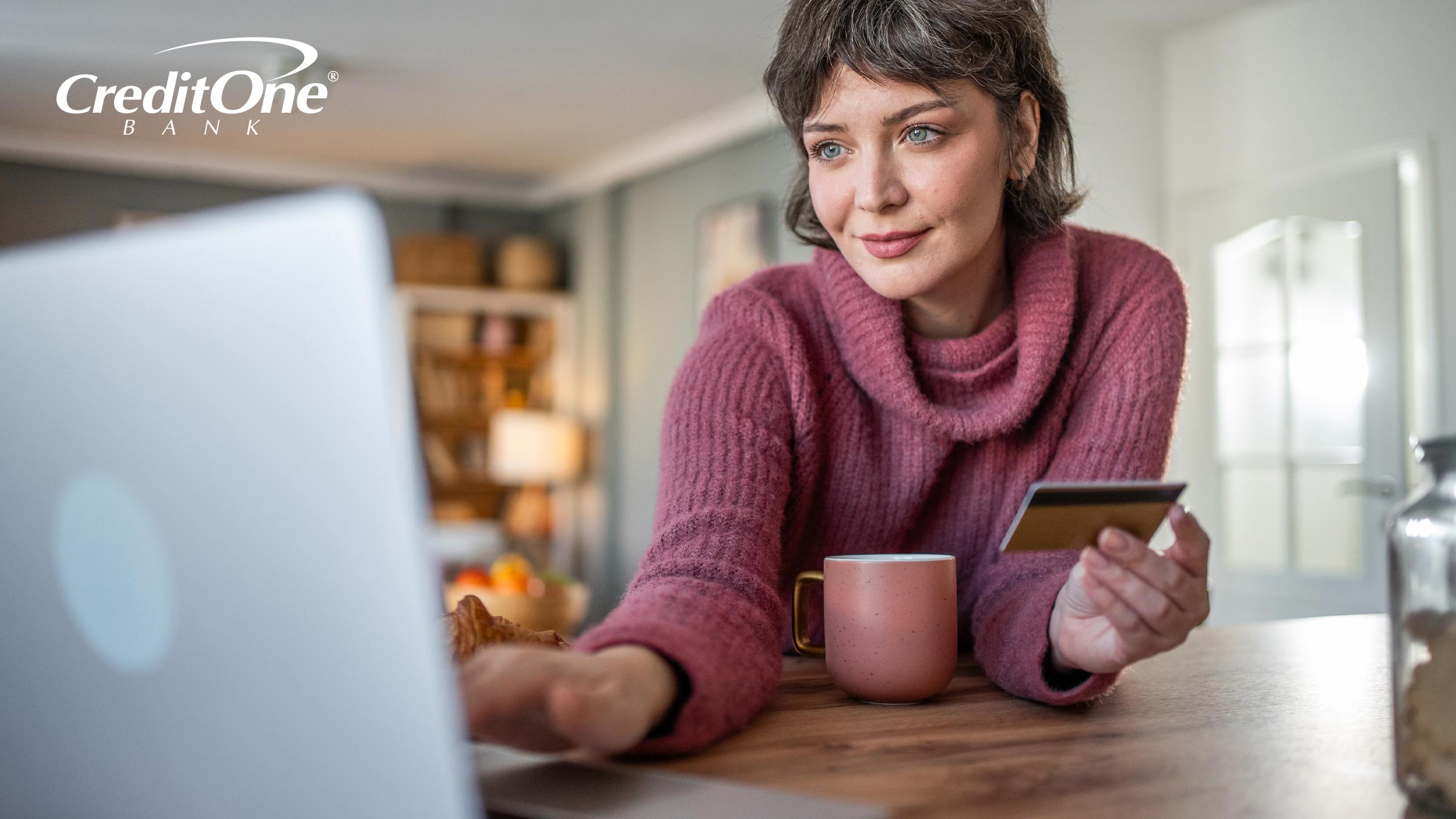 A smiling woman with a plum-colored sweater and matching coffee cup holds her credit card in one hand while using her laptop, possibly managing her credit card account online.
