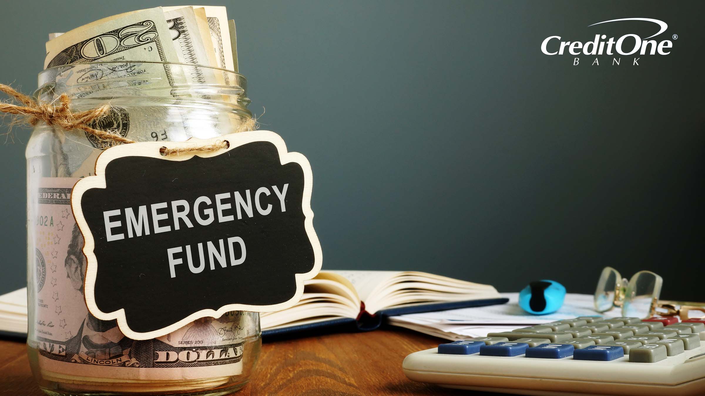 A cash-filled glass jar labeled “Emergency Fund” sits on a desk, representing one of the savings habits consumers might use to prepare for unforeseen expenses.