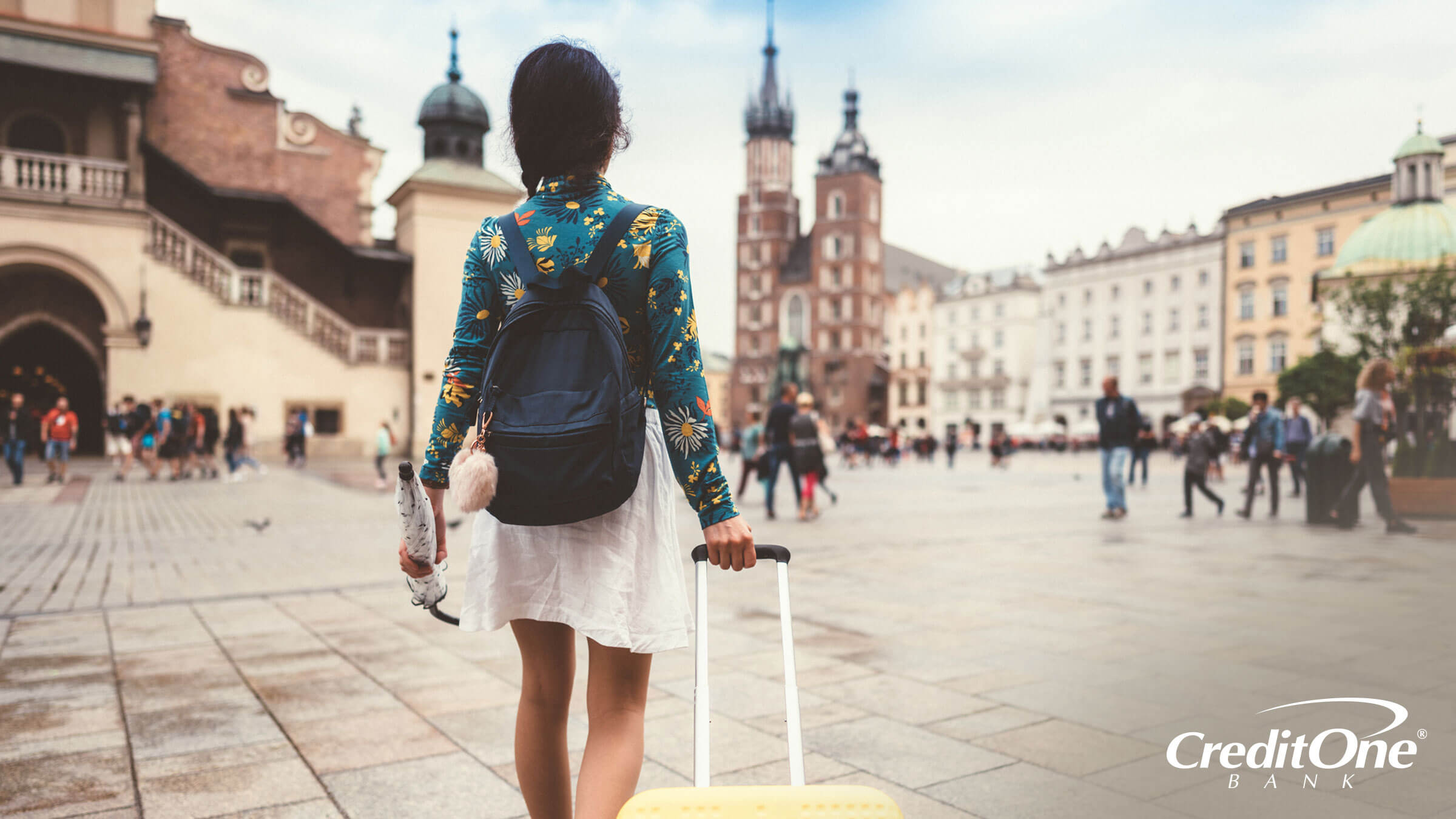 A young woman approaches a large plaza in a European city, with yellow rolling luggage in tow. If she’s studying abroad, she may have used a credit card with rewards to get there.