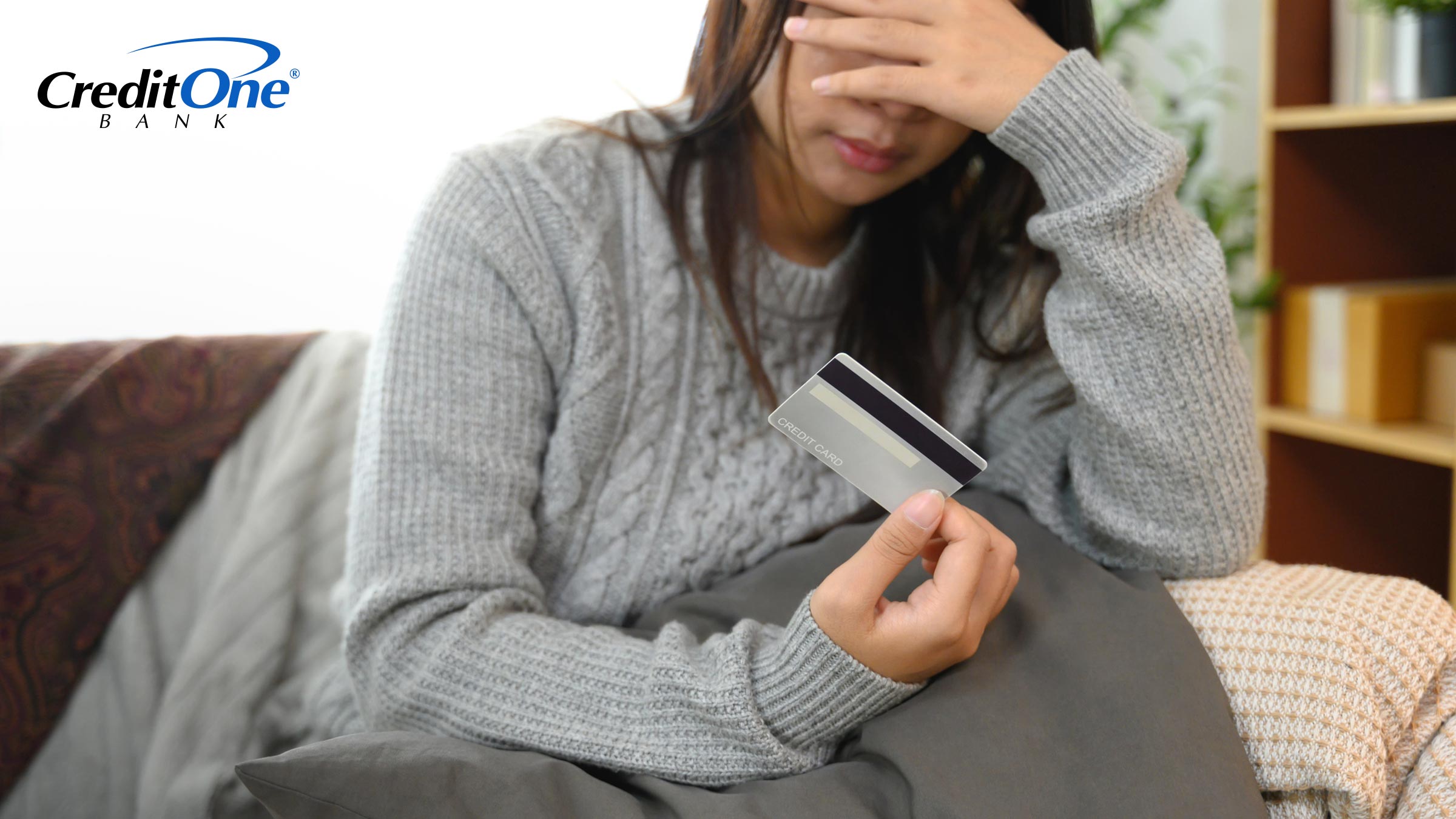 A young woman covers her face while sitting on her couch and holding a credit card in one hand, indicating that she may be confused or stressed about her credit.